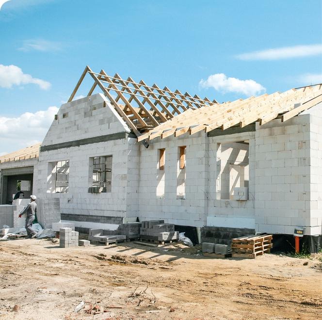 Wznoszenie ścian parterowego domu z białych bloczków, widoczne drewniane elementy konstrukcji dachu, pracownik w oddali.