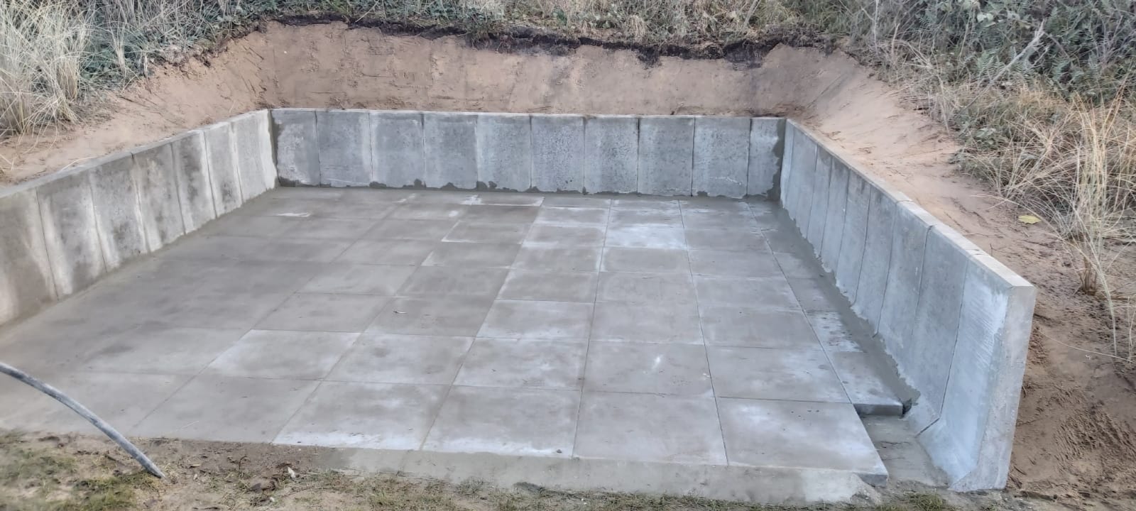 Wykop z betonowymi ścianami oporowymi i wylewką z płyt betonowych na dnie, otoczony ziemią i trawą.