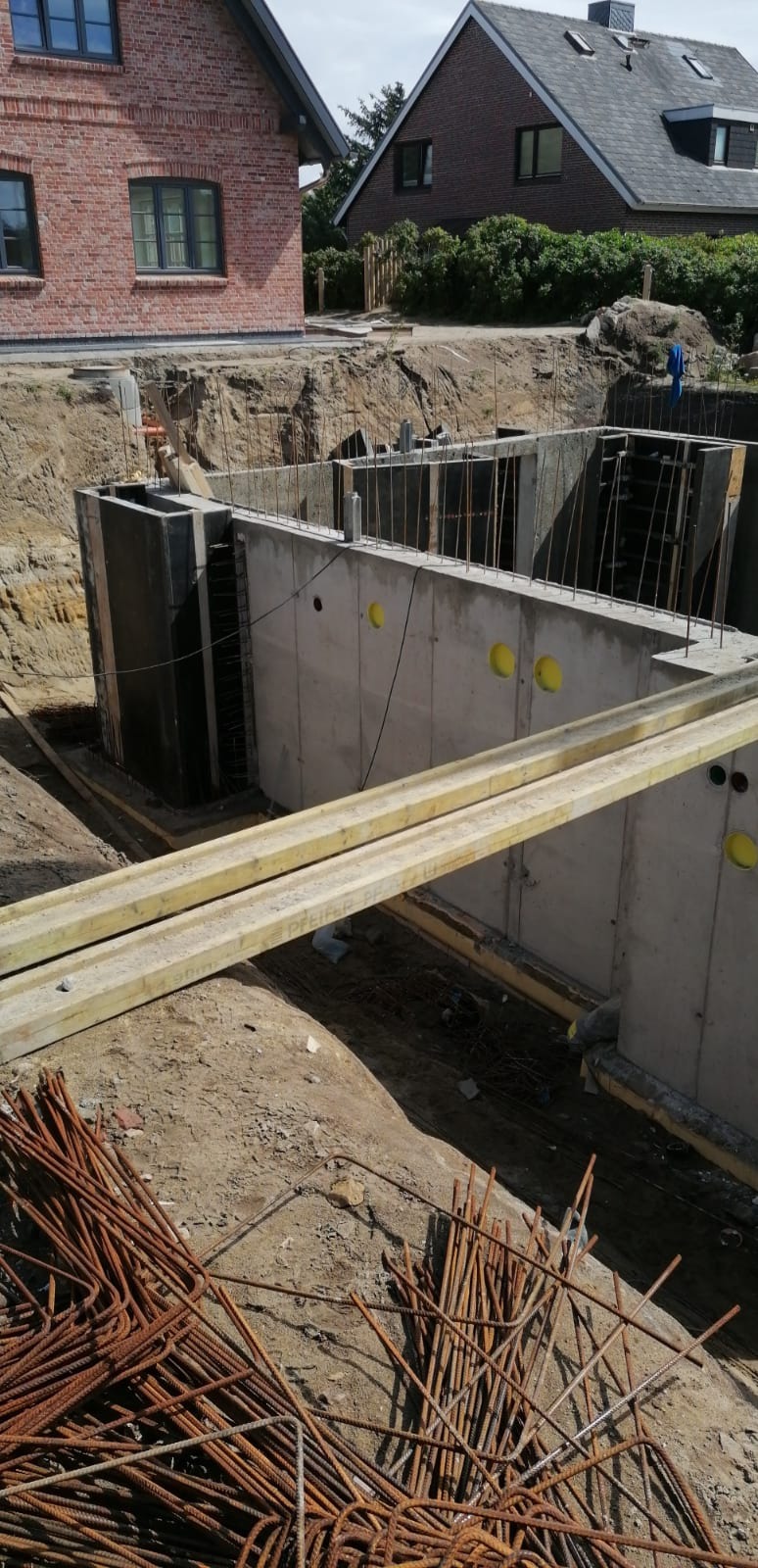 Wykop pod fundamenty domu z wylanym betonem i zbrojeniem, widoczne drewniane stemple podtrzymujące szalunek, w tle budynki mieszkalne.