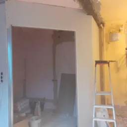 Montaż płyty gipsowo-kartonowej na ścianie, widoczna drabina, materiały budowlane i instalacja elektryczna w tle.