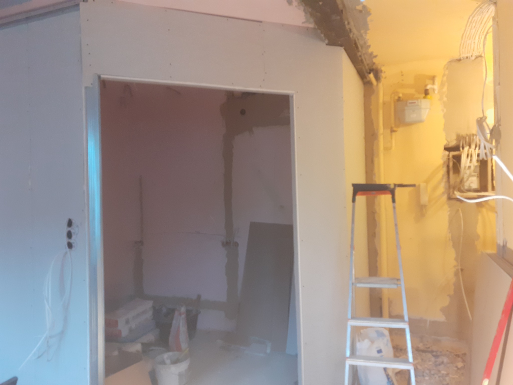 Montaż płyty gipsowo-kartonowej na ścianie, widoczna drabina, materiały budowlane i instalacja elektryczna w tle.