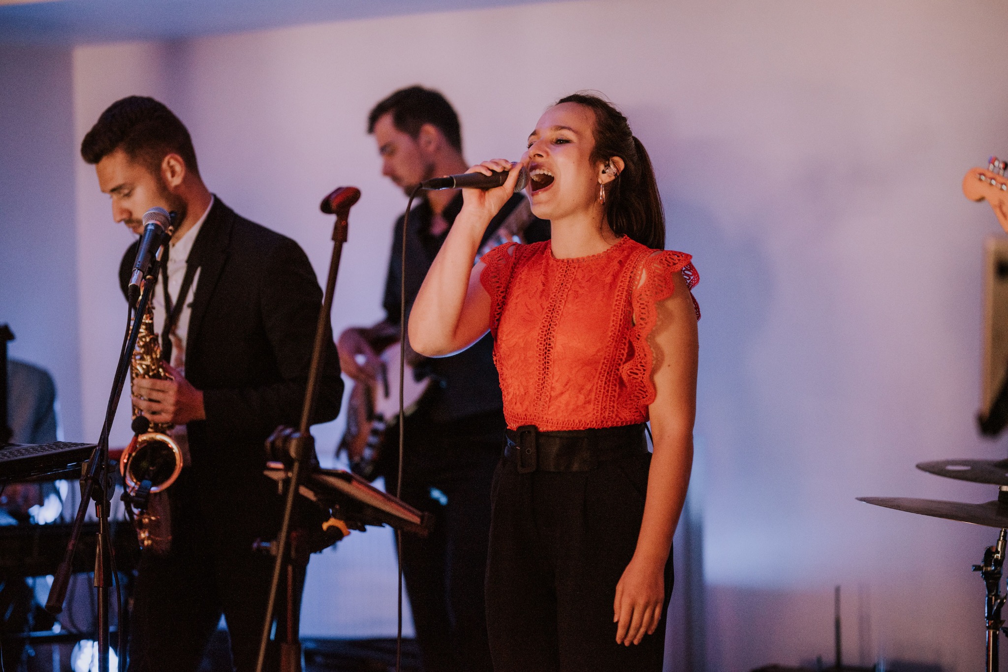 Zespół muzyczny podczas występu: wokalistka w czerwonej bluzce śpiewa do mikrofonu, saksofonista w garniturze i gitarzysta w tle.