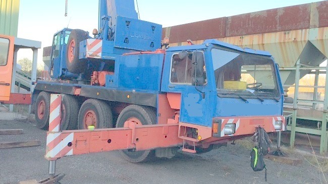 Niebiesko-pomarańczowy dźwig samochodowy Liebherr na placu budowy, z widocznym wysięgnikiem i podporami, obok konstrukcji magazynowej.