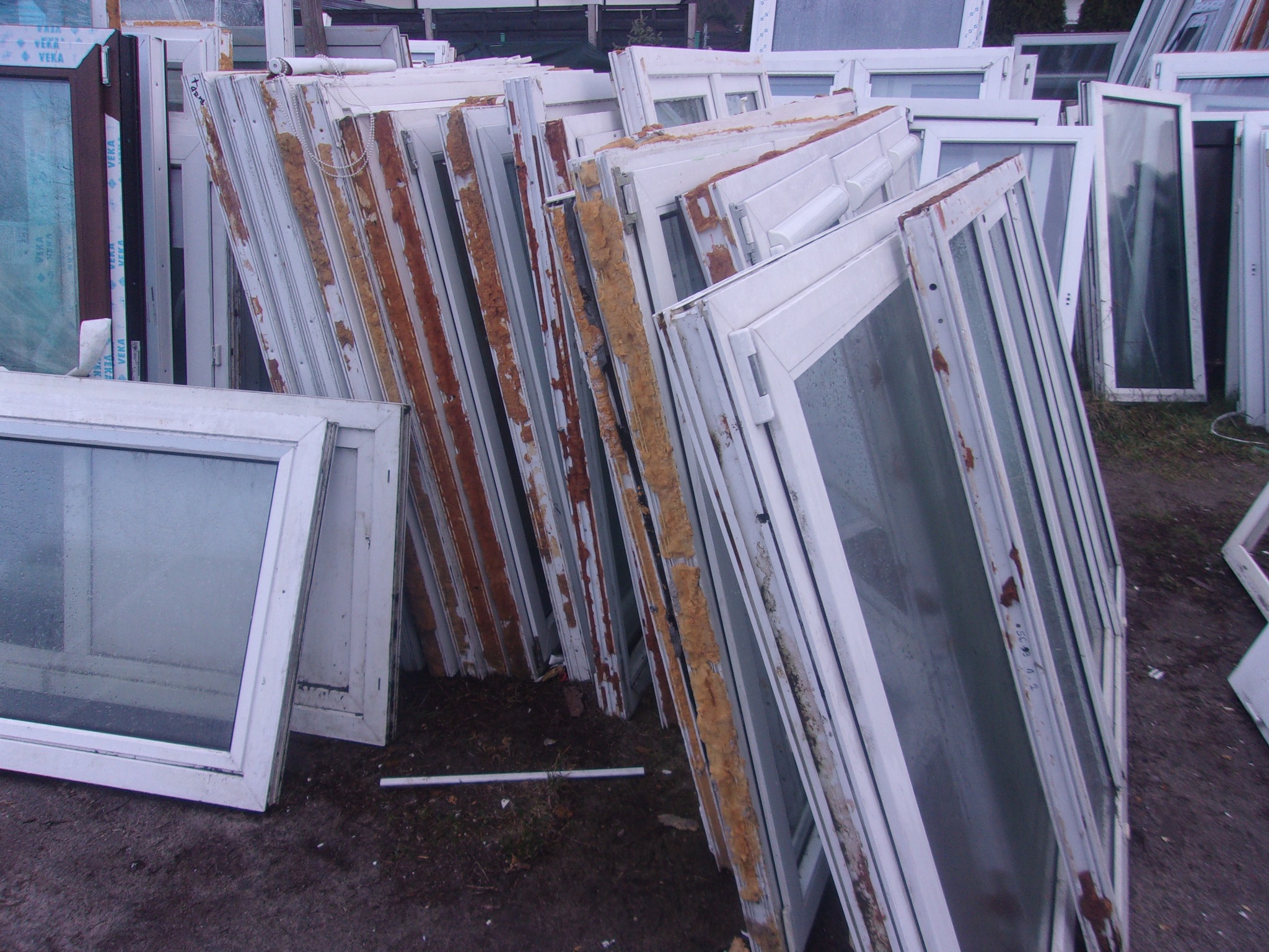 Stos białych ram okiennych z widocznymi śladami demontażu, brudne szyby, składowane na zewnątrz.