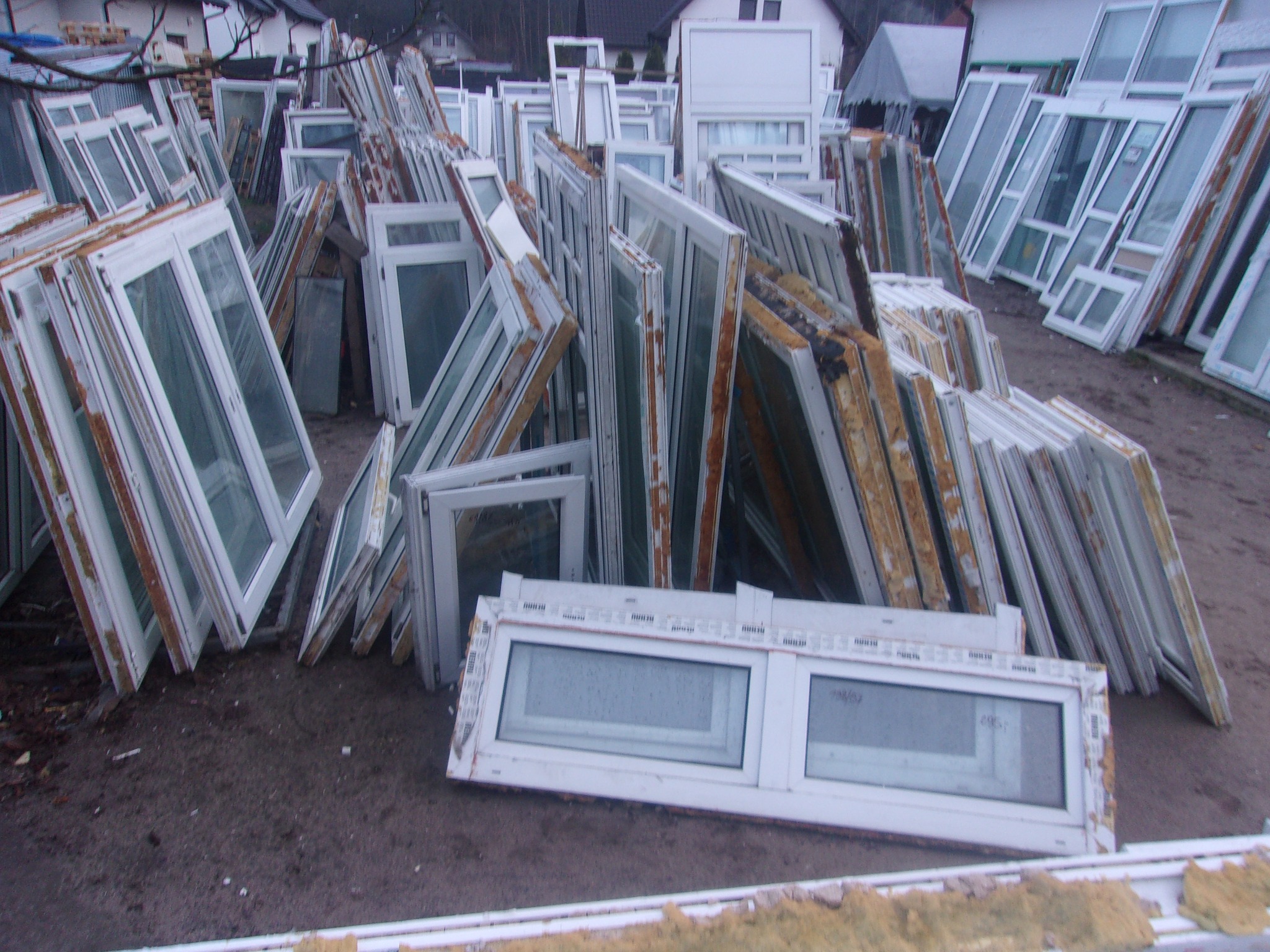 Stos starych okien PCV i aluminiowych, niektóre z widocznymi śladami użytkowania i uszkodzeniami, składowane na zewnątrz, w tle budynki mieszkalne.