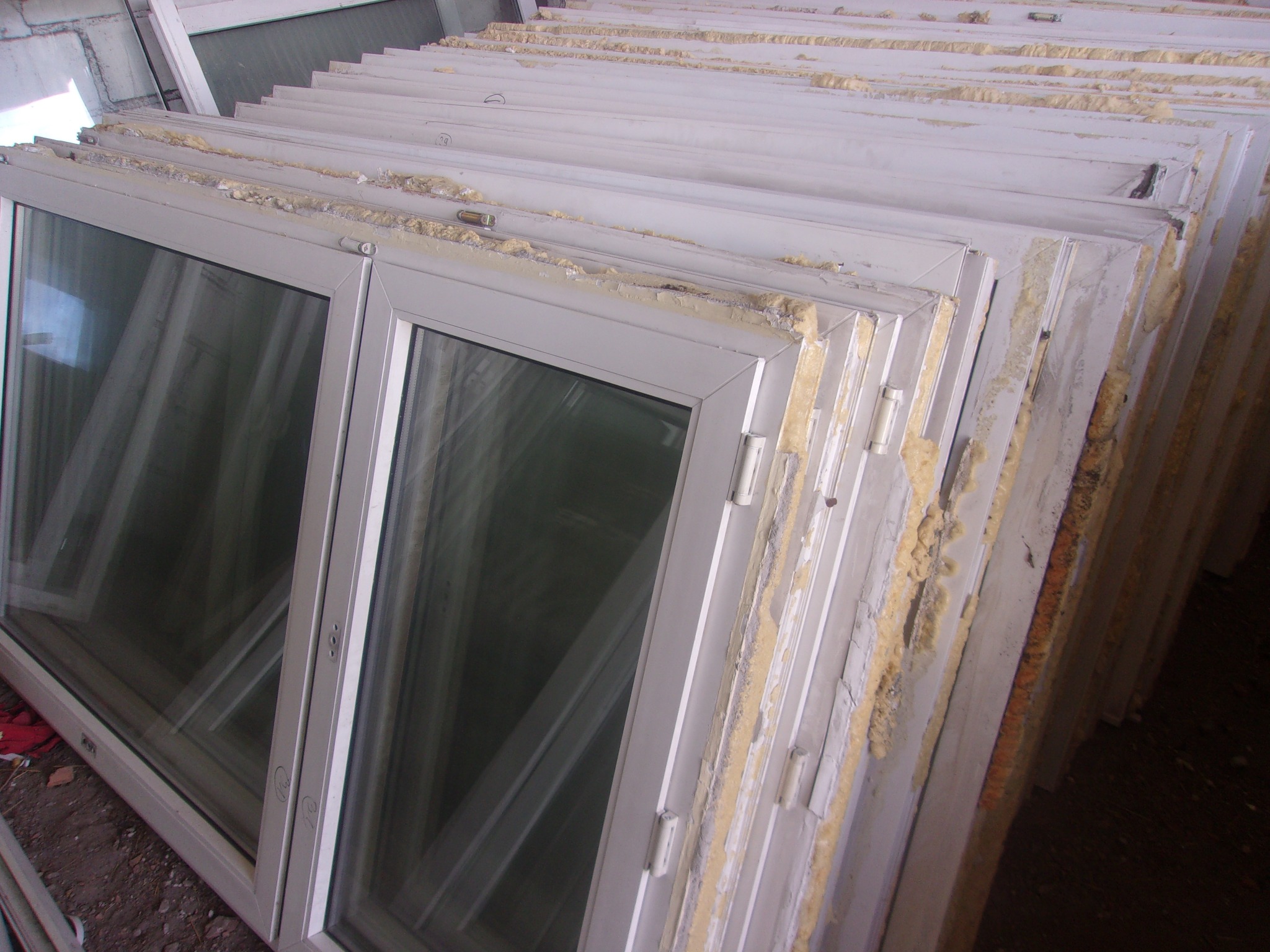 Stos białych okien przygotowanych do montażu, widoczne resztki pianki montażowej i oznaczenia na ramach.