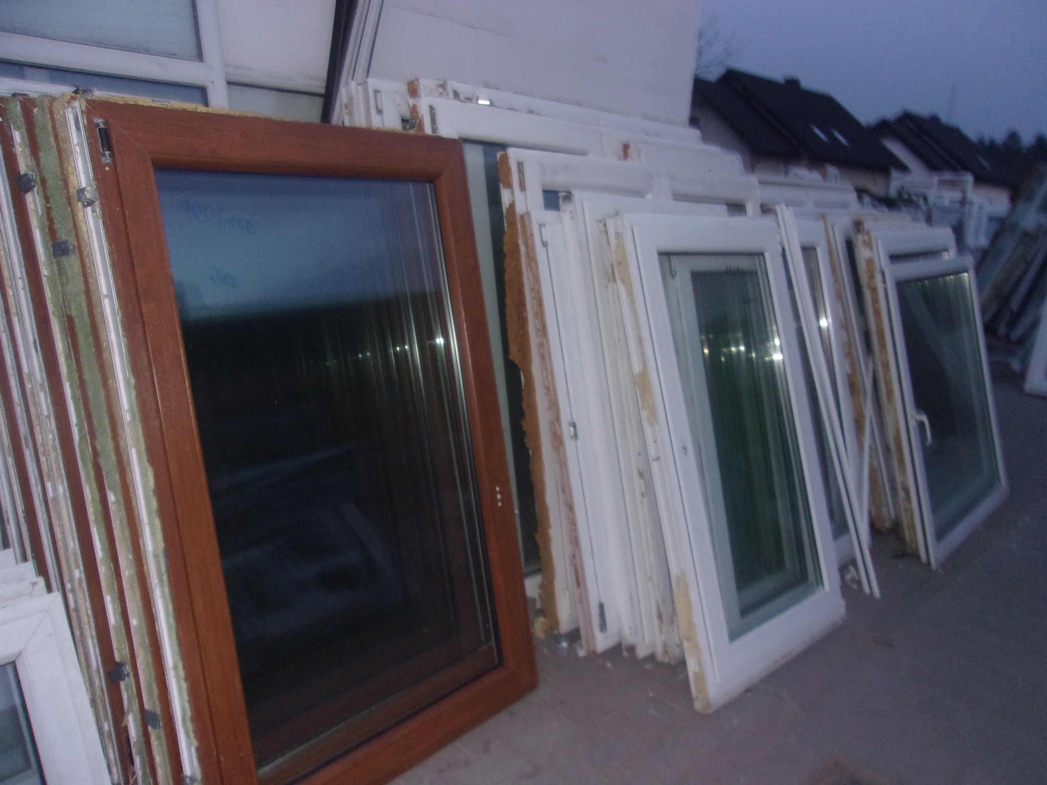 Magazyn okien: brązowa rama okienna na pierwszym planie, za nią rząd białych okien opartych o siebie, widoczne ślady pianki montażowej i zabrudzenia.