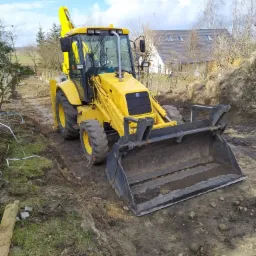 Żółta koparko-ładowarka na placu budowy z wytyczonym terenem, widoczne drzewa i dom w tle.