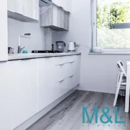 Jasna, minimalistyczna kuchnia z białymi, błyszczącymi frontami szafek, szarymi drewnianymi akcentami w górnych szafkach i blatem, widok na okno i krzesło w tle, jasna podłoga imitująca drewno.