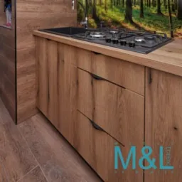 Blat kuchenny z drewna o wyraźnym usłojeniu, czarny zlew i płyta gazowa, fronty szafek z podłużnymi, czarnymi uchwytami, fototapeta z motywem leśnym w tle, widoczne logo firmy w prawym dolnym rogu.