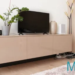 Beżowa szafka RTV wisząca z połyskiem, z czarnym cokołem, na której stoi telewizor, głośniki, roślina w doniczce i dekoracyjny wazon, z widocznymi kablami pod spodem.