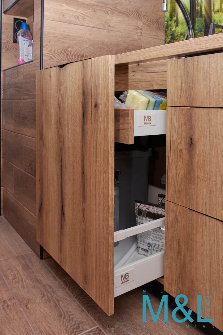 Otwarta szafka kuchenna z drewnopodobnym frontem, wysuwane półki z koszem na śmieci i akcesoriami do sprzątania, widoczna butelka z płynem w górnej szafce.