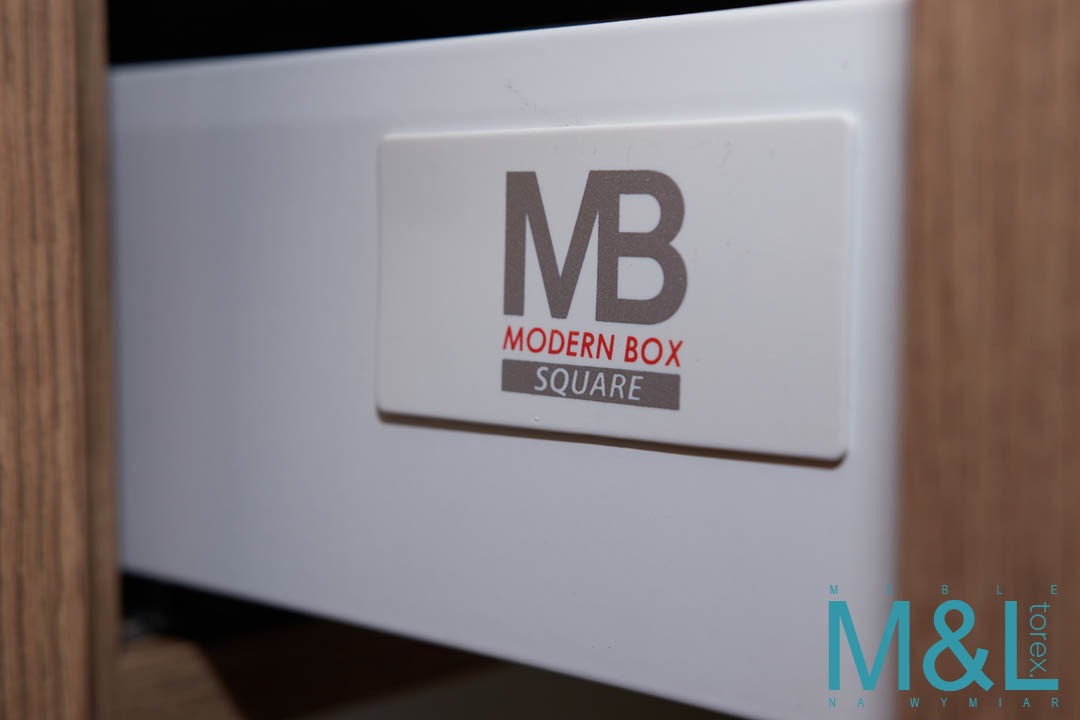 Biała szuflada z systemem Modern Box Square, widoczna etykieta producenta i fragment drewnianego frontu meblowego.