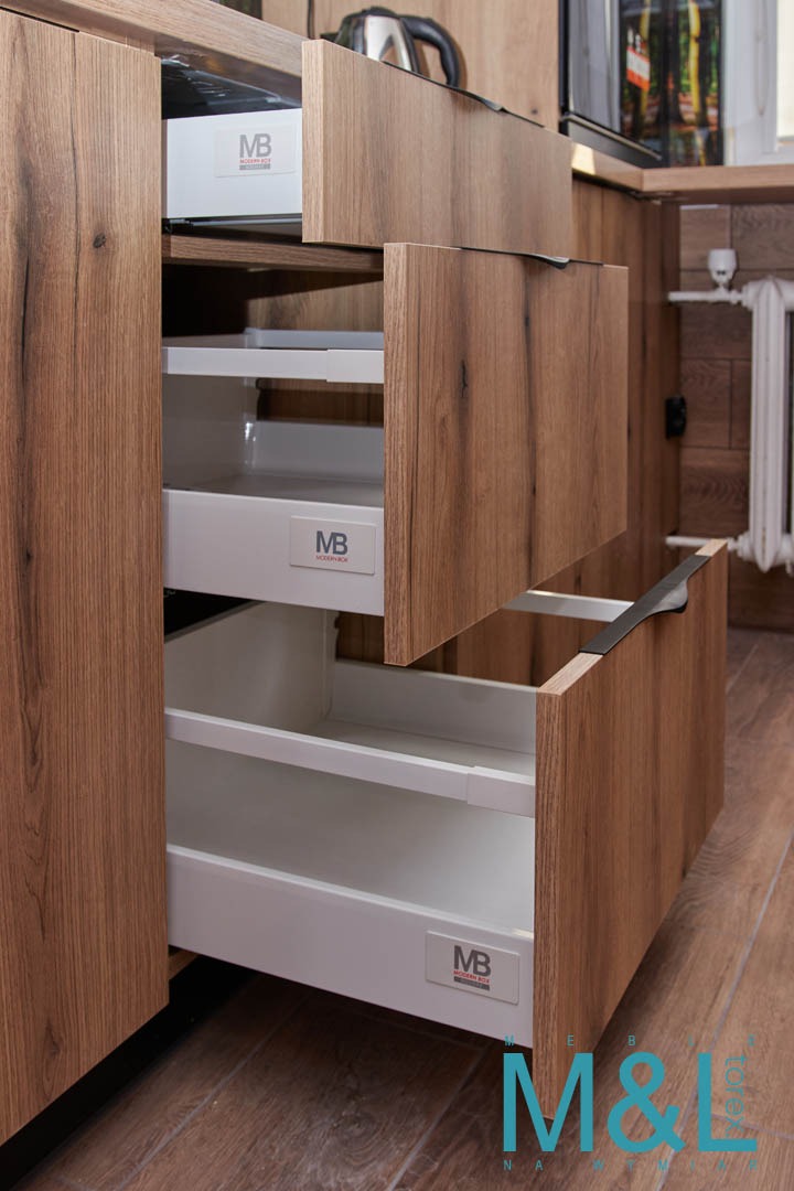 Wysuwane szuflady z drewnianymi frontami w kuchni, widoczne białe wnętrza szuflad z logo producenta, czarne uchwyty, parkiet na podłodze.