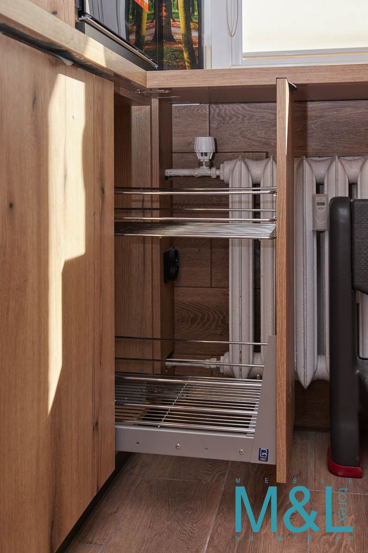 Wysuwana metalowa półka w szafce kuchennej z drewnianymi frontami, widoczny grzejnik za szafką i logo firmy na dolnej krawędzi półki.