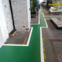 Świeżo pomalowana na zielono podłoga w hali przemysłowej z wyznaczonymi białymi pasami, tworząca ciąg komunikacyjny, obok kolumny z cegły pomalowanej na biało.