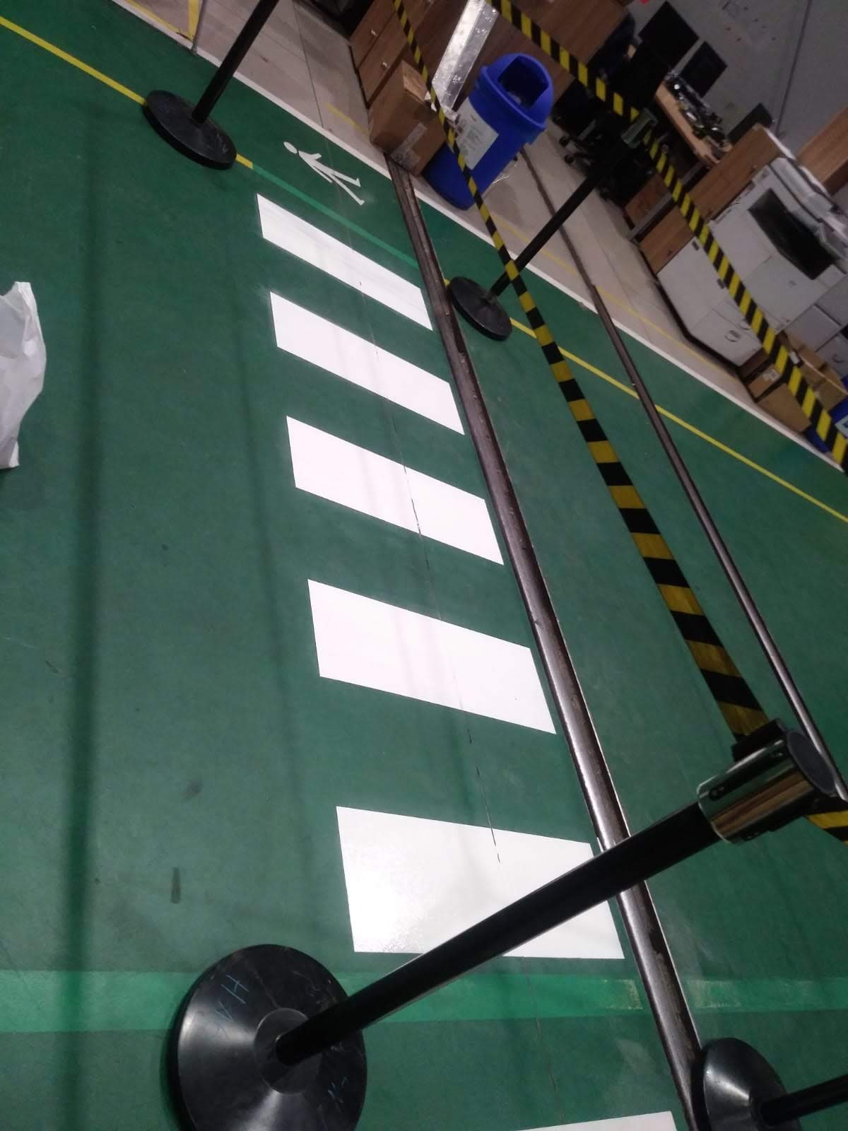 Białe pasy przejścia dla pieszych i symbol osoby na zielonej podłodze hali produkcyjnej, wyznaczające bezpieczną strefę, z widocznymi słupkami oddzielającymi przestrzeń.