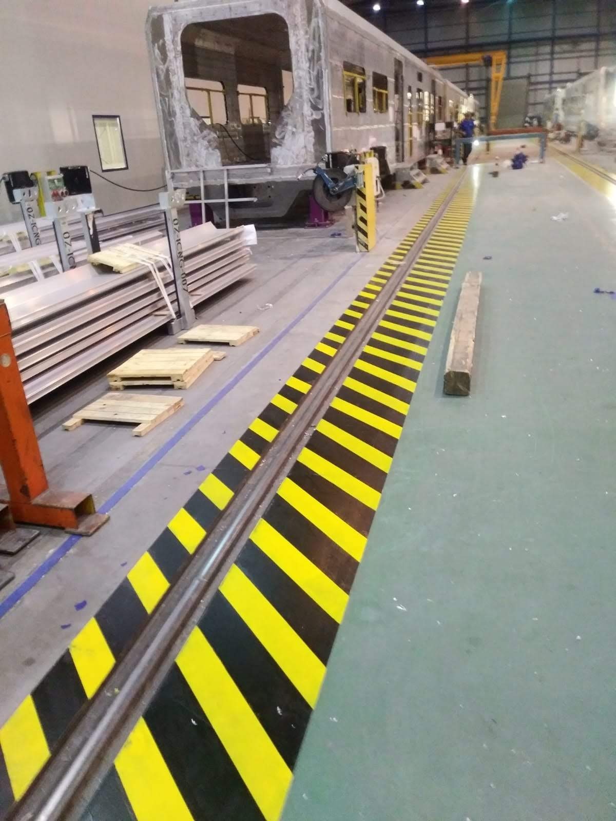 Wnętrze hali produkcyjnej w Gliwicach z surowym szkieletem wagonu kolejowego w trakcie budowy lub naprawy, widoczne elementy konstrukcyjne, podłoga z pasami ostrzegawczymi i stos blach.