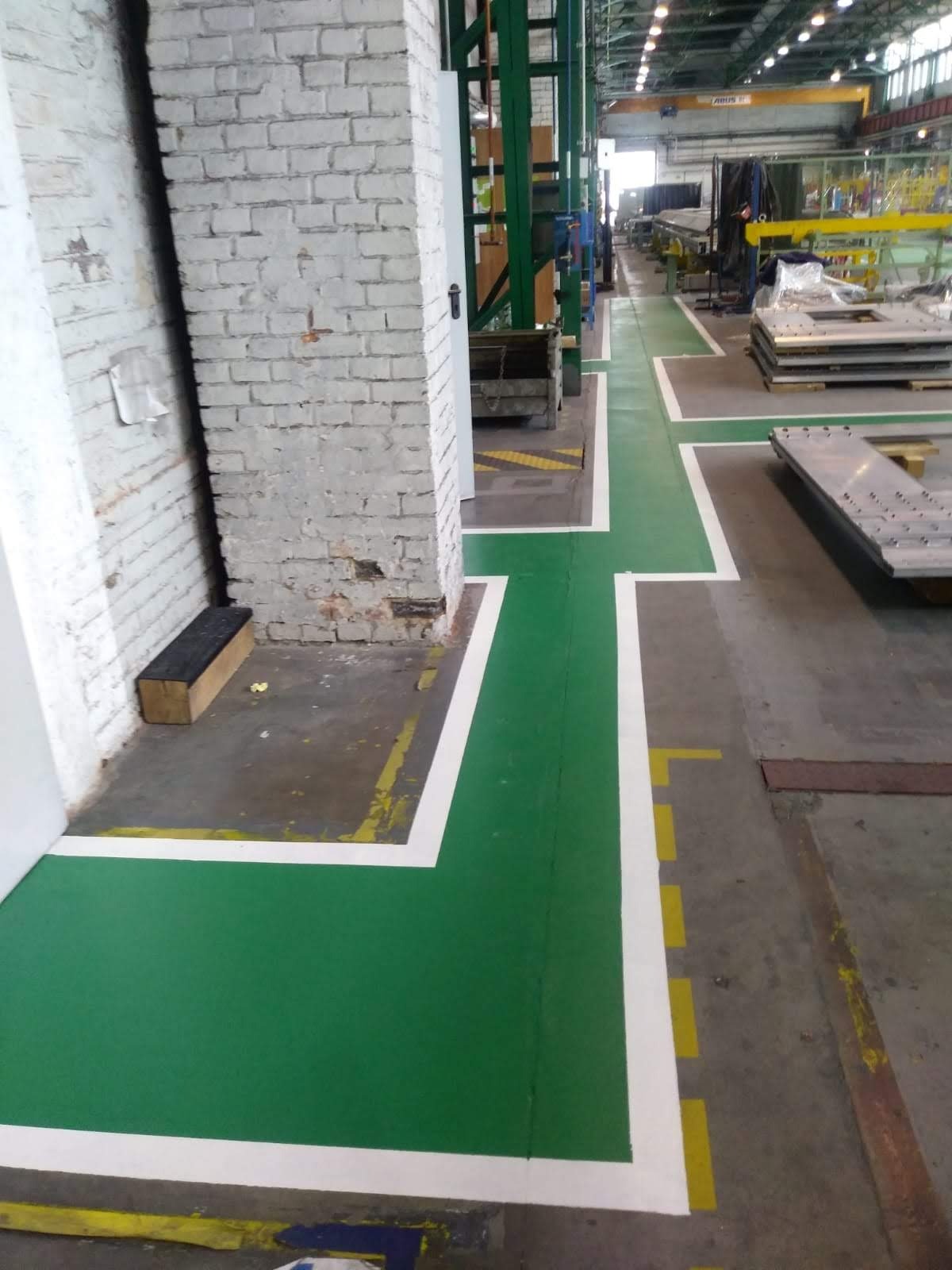 Świeżo pomalowana na zielono podłoga w hali przemysłowej z wyznaczonymi białymi pasami, tworząca ciąg komunikacyjny, obok kolumny z cegły pomalowanej na biało.