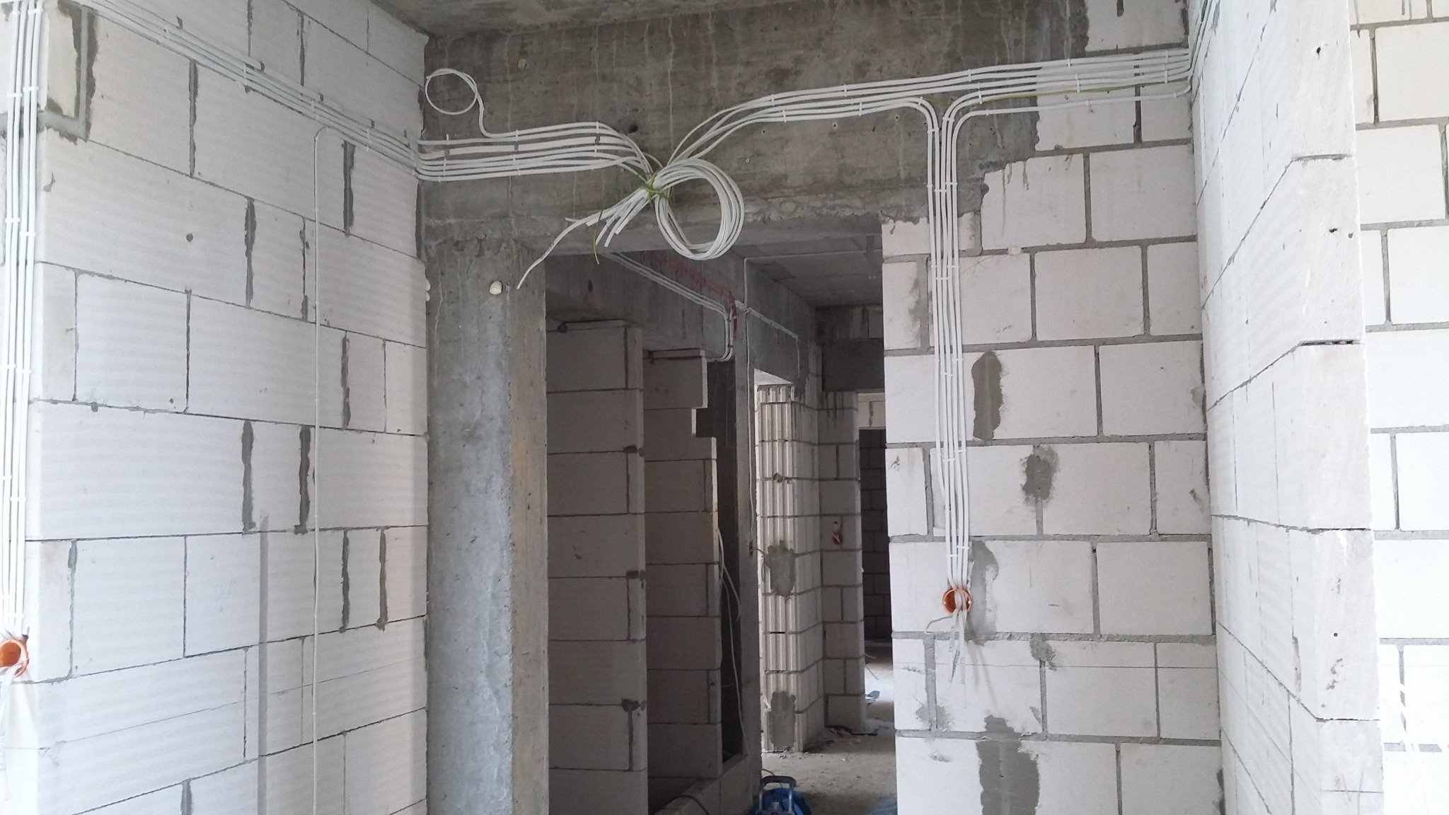 Białe kable elektryczne prowadzone po ścianach z betonu komórkowego w surowym stanie, z widocznymi puszkami instalacyjnymi i zwisającym pęczkiem kabli, w trakcie instalacji.