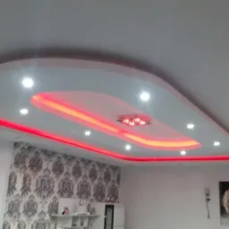 Sufit podwieszany z oświetleniem LED w kolorze czerwonym, centralny element dekoracyjny z żarówkami, wzorzysta tapeta na ścianie w tle, zegar i obraz na ścianie.