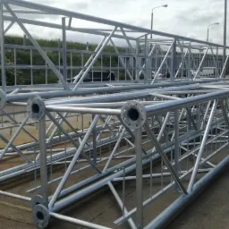 Dwie ocynkowane kratownice stalowe z okrągłymi kołnierzami na końcach, leżące na betonowej powierzchni w otoczeniu zieleni i budynków przemysłowych.