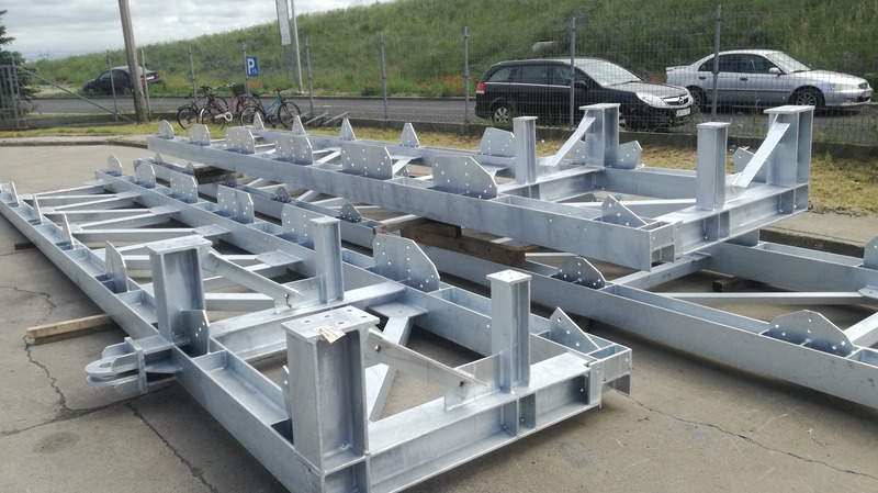 Dwie ocynkowane, spawane ramy konstrukcji stalowej z licznymi otworami na śruby, leżące na betonowym podłożu, z widocznymi samochodami i zielenią w tle.