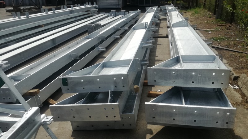 Srebrne, ocynkowane profile stalowe w kształcie litery U, przygotowane do budowy hali, z widocznymi otworami na śruby i spawami, składowane na zewnątrz.