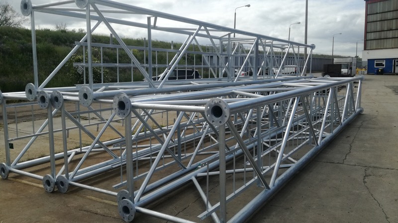 Dwie ocynkowane kratownice stalowe z okrągłymi kołnierzami na końcach, leżące na betonowej powierzchni w otoczeniu zieleni i budynków przemysłowych.