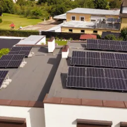 Instalacja fotowoltaiczna na płaskim dachu budynku mieszkalnego, widok z góry, z zielonym otoczeniem i zabudowaniami w tle.