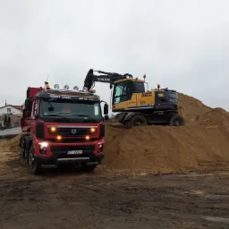 Czerwona ciężarówka Volvo z napisem 'ROBOTY ZIEMNE' na tle żółtej koparki Volvo EW140C stojącej na usypanej górze piasku na placu budowy.