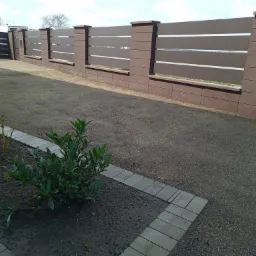 Nowoczesne ogrodzenie panelowe z bramą wjazdową, brązowe słupki z betonowych bloczków, trawnik z młodymi roślinami i ścieżką z kostki.