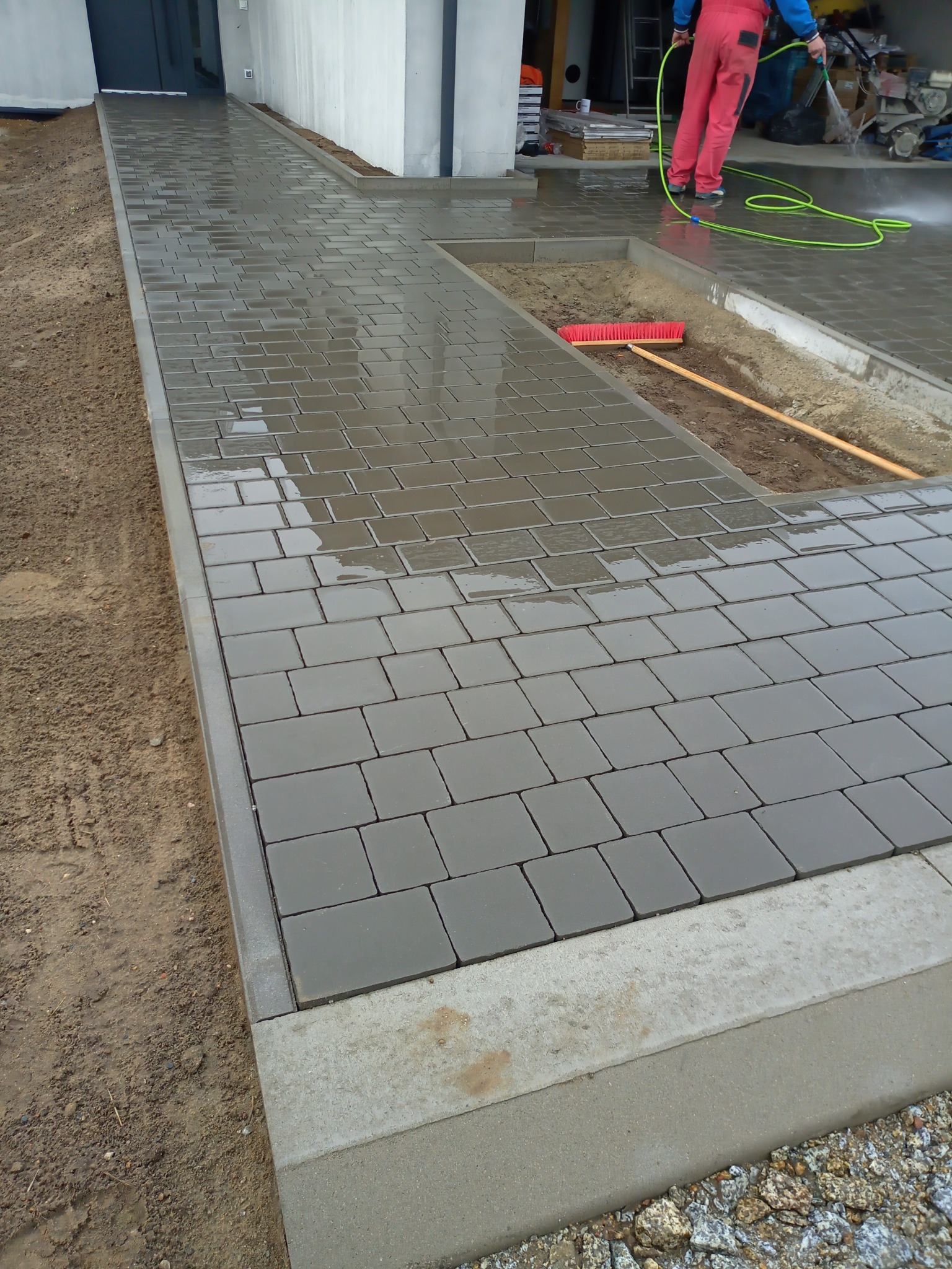 Świeżo ułożona, mokra kostka brukowa w odcieniach szarości, obramowana betonowym krawężnikiem, z widocznym fragmentem ziemi i robotnikiem czyszczącym powierzchnię za pomocą węża z wodą.