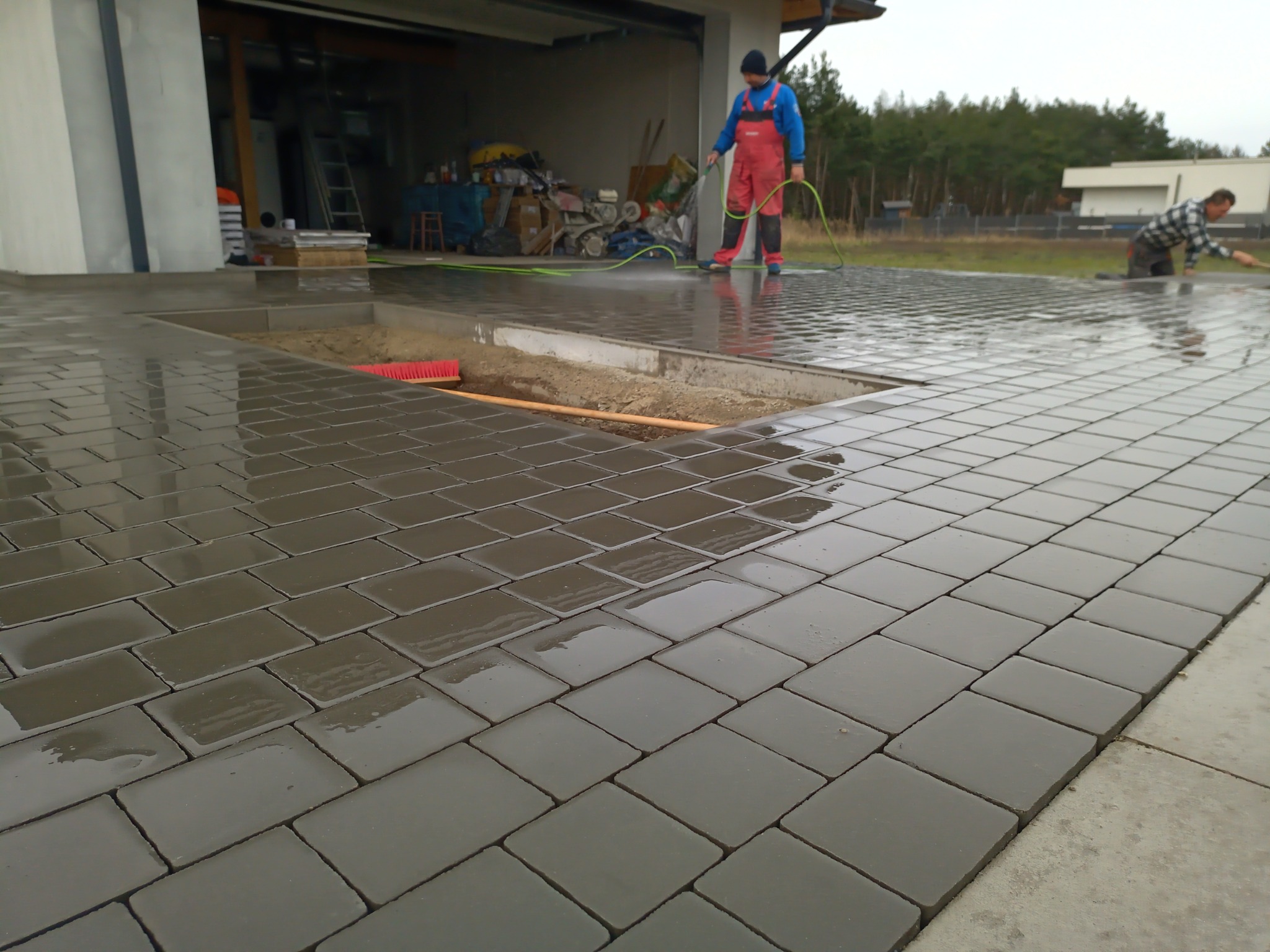 Dwaj pracownicy czyszczą wodą nowo ułożoną, mokrą, szarą kostkę brukową na podjeździe przed garażem, z widocznym fragmentem koryta z podsypką i czerwoną szczotką.