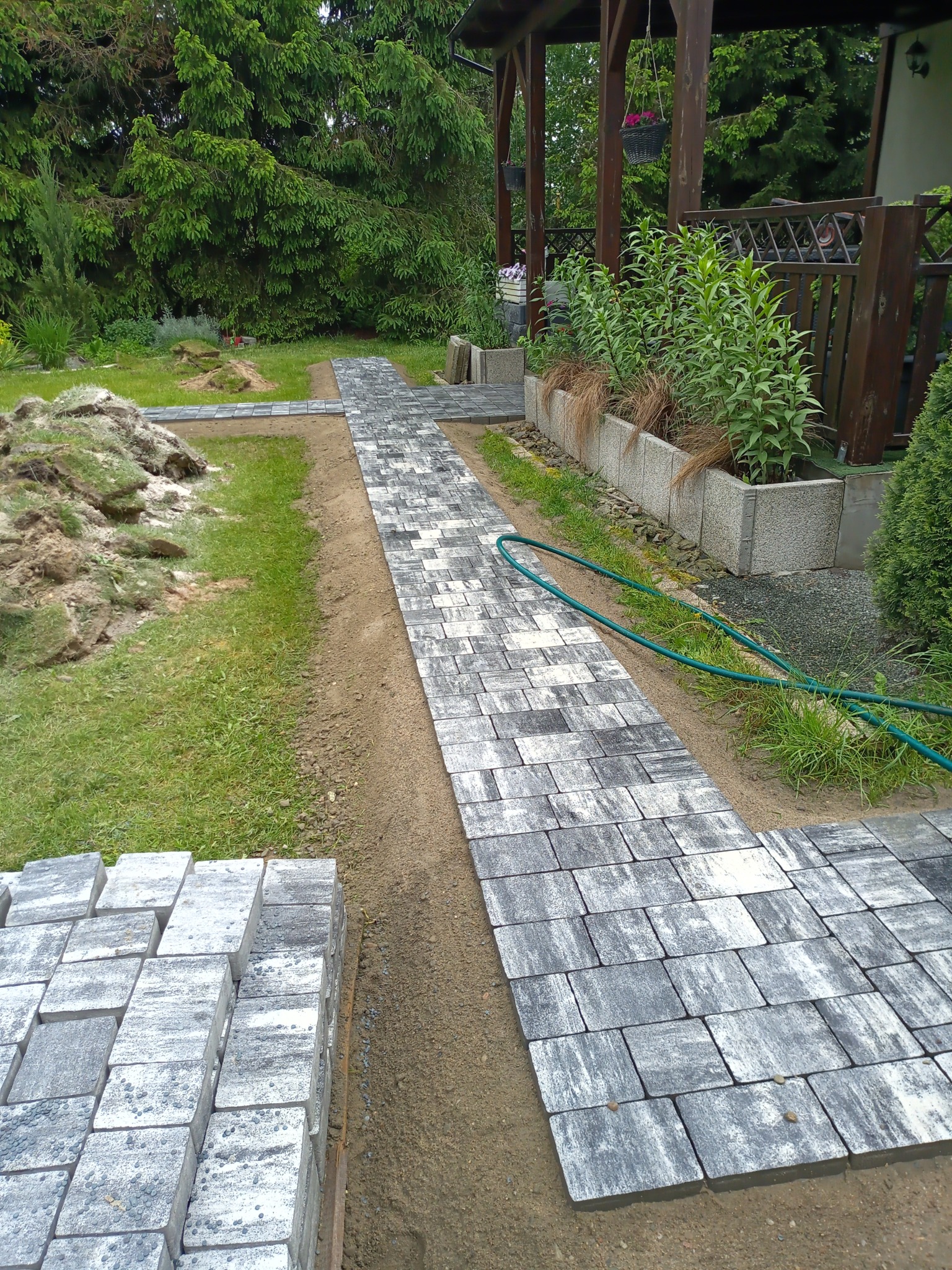 Układanie szarej kostki brukowej w ogrodzie, widoczny fragment ścieżki i stos kostki do dalszej pracy, trawnik z wykopaną ziemią w tle.
