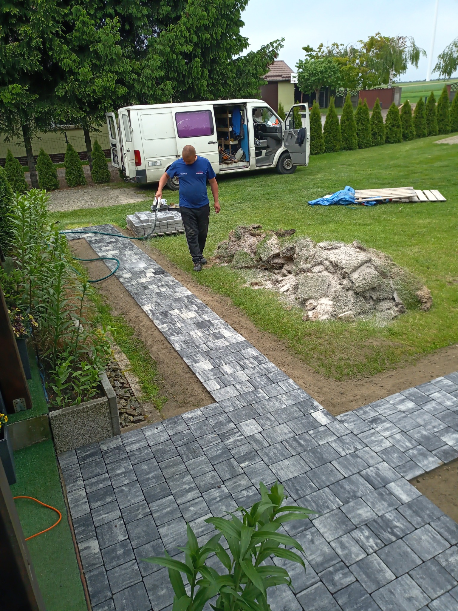 Mężczyzna w niebieskiej koszulce, z wężem w ręku, nawilża świeżo ułożoną ścieżkę z szarej kostki brukowej na posesji; w tle biały samochód dostawczy z otwartymi drzwiami, sterta zdjętej darni...