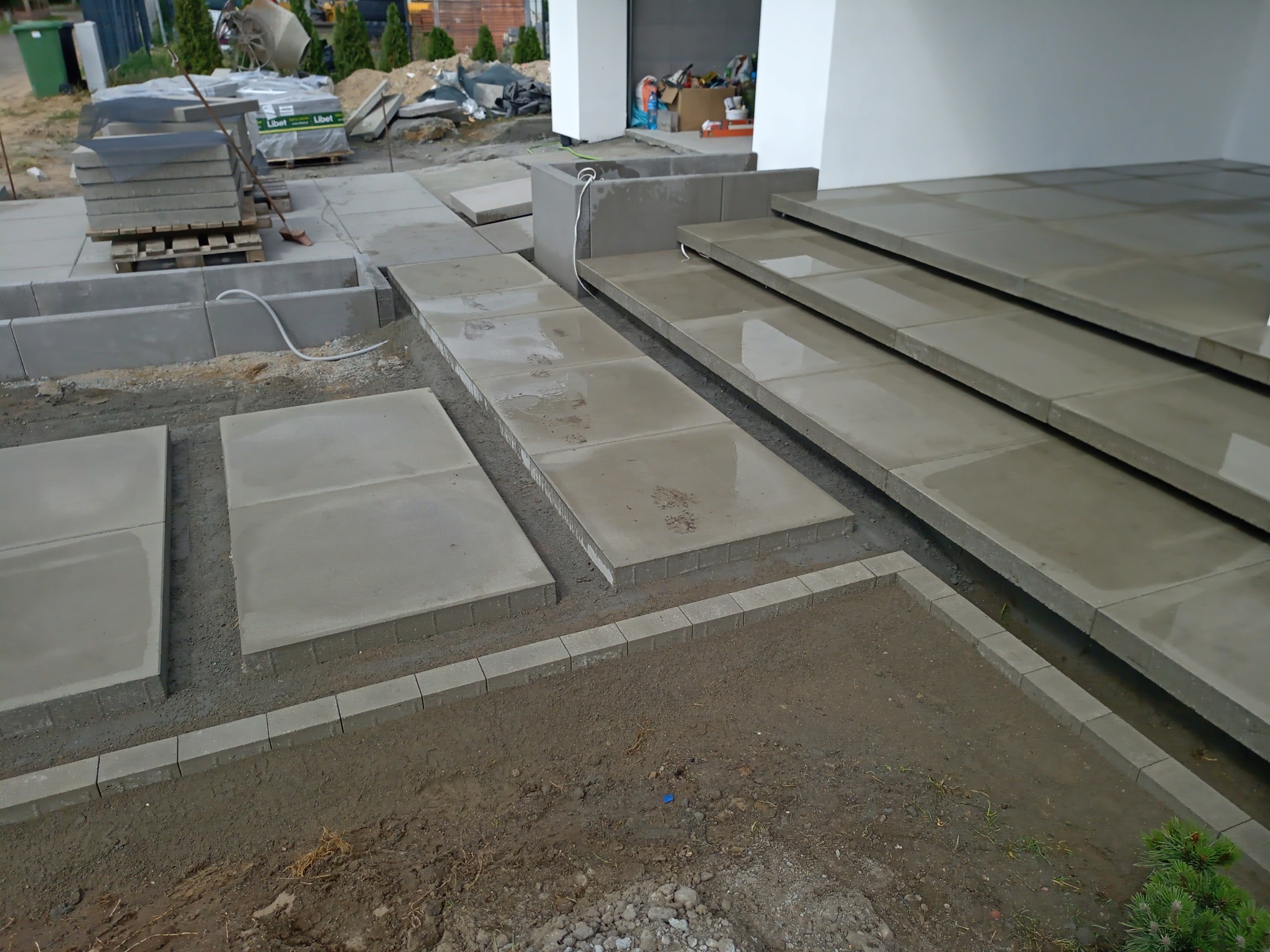 Układanie dużych, szarych płyt chodnikowych i obrzeży z kostki na podwórku, widoczne palety z kostką w tle.