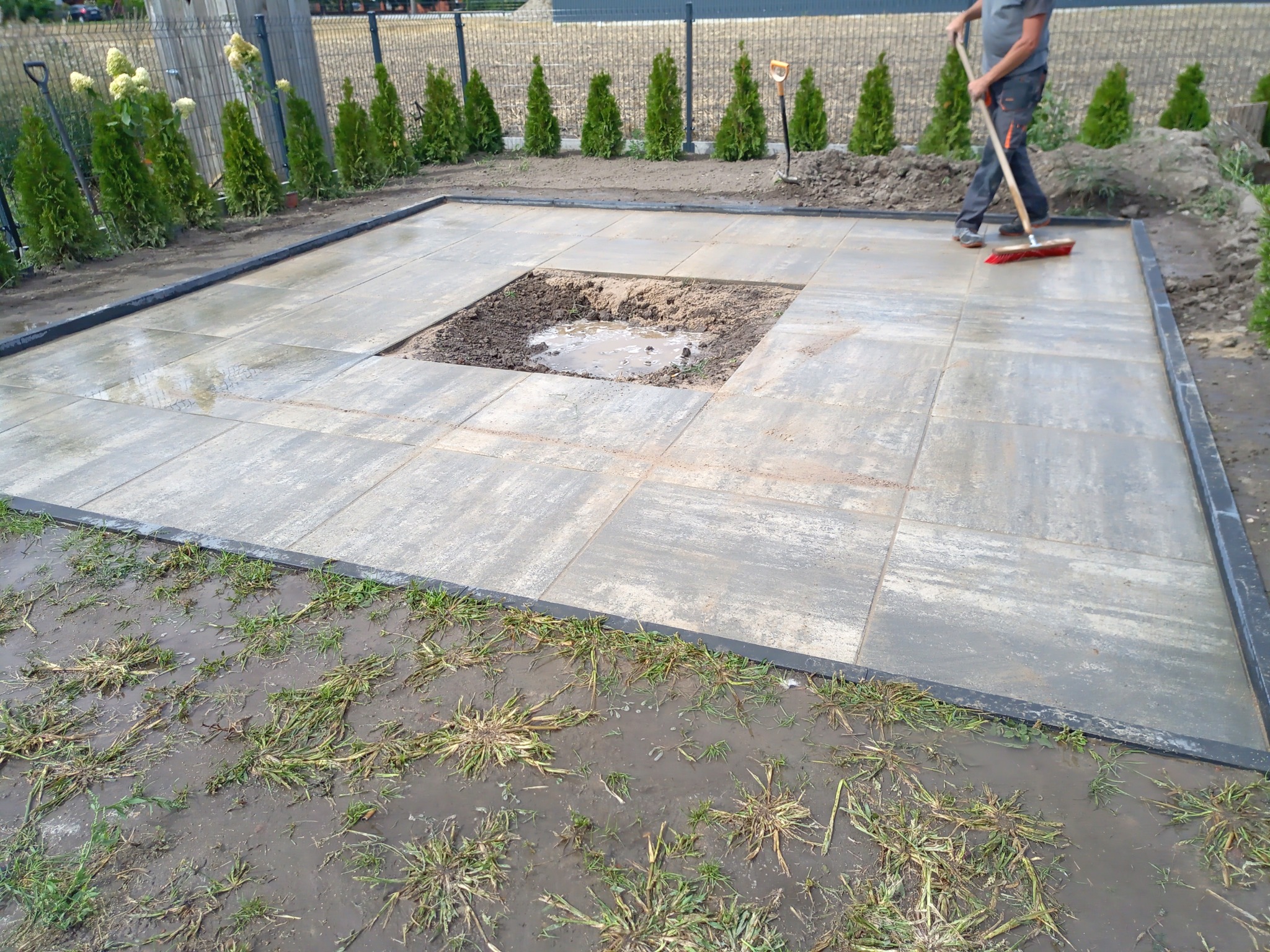 Układanie szarej kostki brukowej na przygotowanym podłożu, z wykopanym kwadratem w środku, pracownik zamiata powierzchnię, widoczne rośliny w tle.