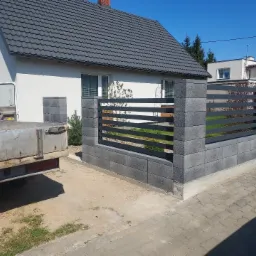 Nowoczesne ogrodzenie z szarej kostki betonowej z poziomymi, metalowymi przęsłami, częściowo ukończone; widoczny samochód dostawczy z tyłu