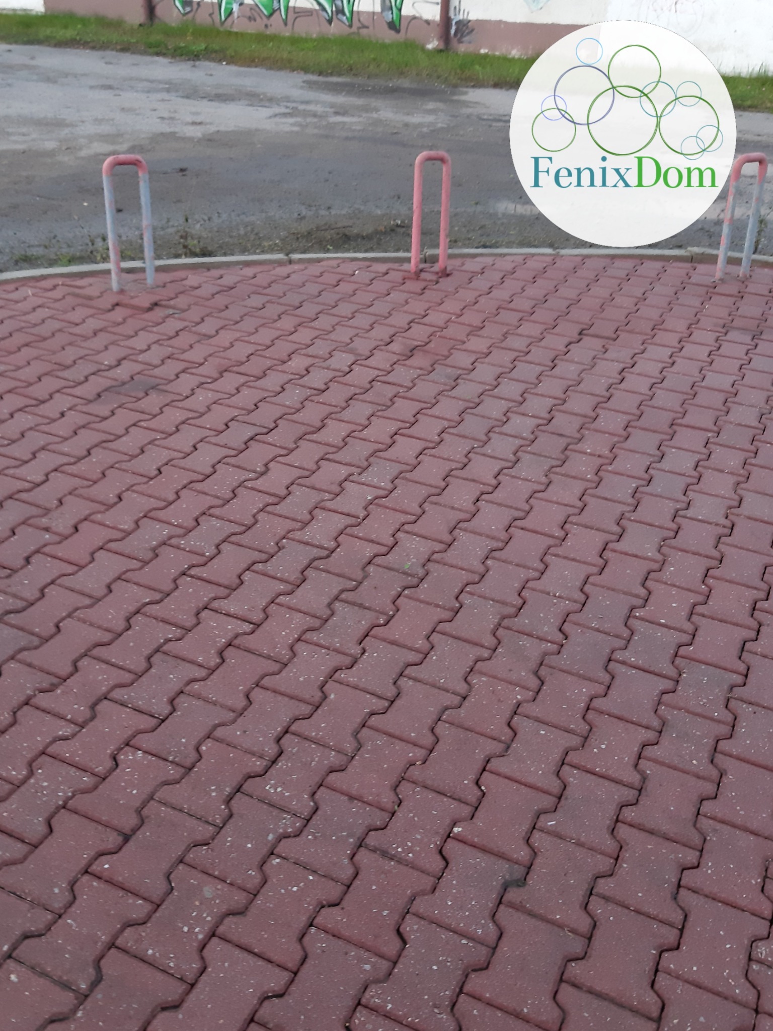 Czerwona kostka brukowa ułożona w jodełkę, z widocznymi fragmentami metalowych słupków i logo firmy FenixDom w górnym rogu.