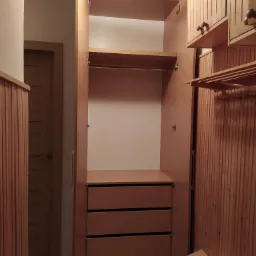 Wnętrze garderoby z drewnianą zabudową: szafa z trzema szufladami, półkami i drążkiem na ubrania, widoczne pionowe panele na ścianie i drzwiach w tle.