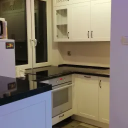 Biała kuchnia z czarnym blatem, górne szafki z ażurowymi frontami, oświetlenie punktowe, podłoga w odcieniach brązu ułożona w jodełkę.