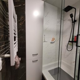 Mookodi - Nowoczesna łazienka z kabiną prysznicową typu walk-in, białą szafką z czarnymi uchwytami i grzejnikiem drabinkowym na tle marmurowej ściany.