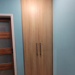 Mookodi - Jasna, drewniana szafa dwudrzwiowa z długimi, czarnymi uchwytami, wkomponowana we wnękę pokoju z błękitnymi ścianami i drewnianą podłogą. Minimalistyczny design.