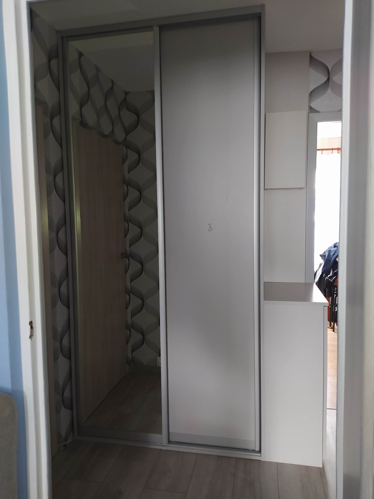 Biała szafa wnękowa z przesuwnymi drzwiami, widoczna w korytarzu z wzorzystą tapetą, częściowo otwarta ukazująca wnętrze.