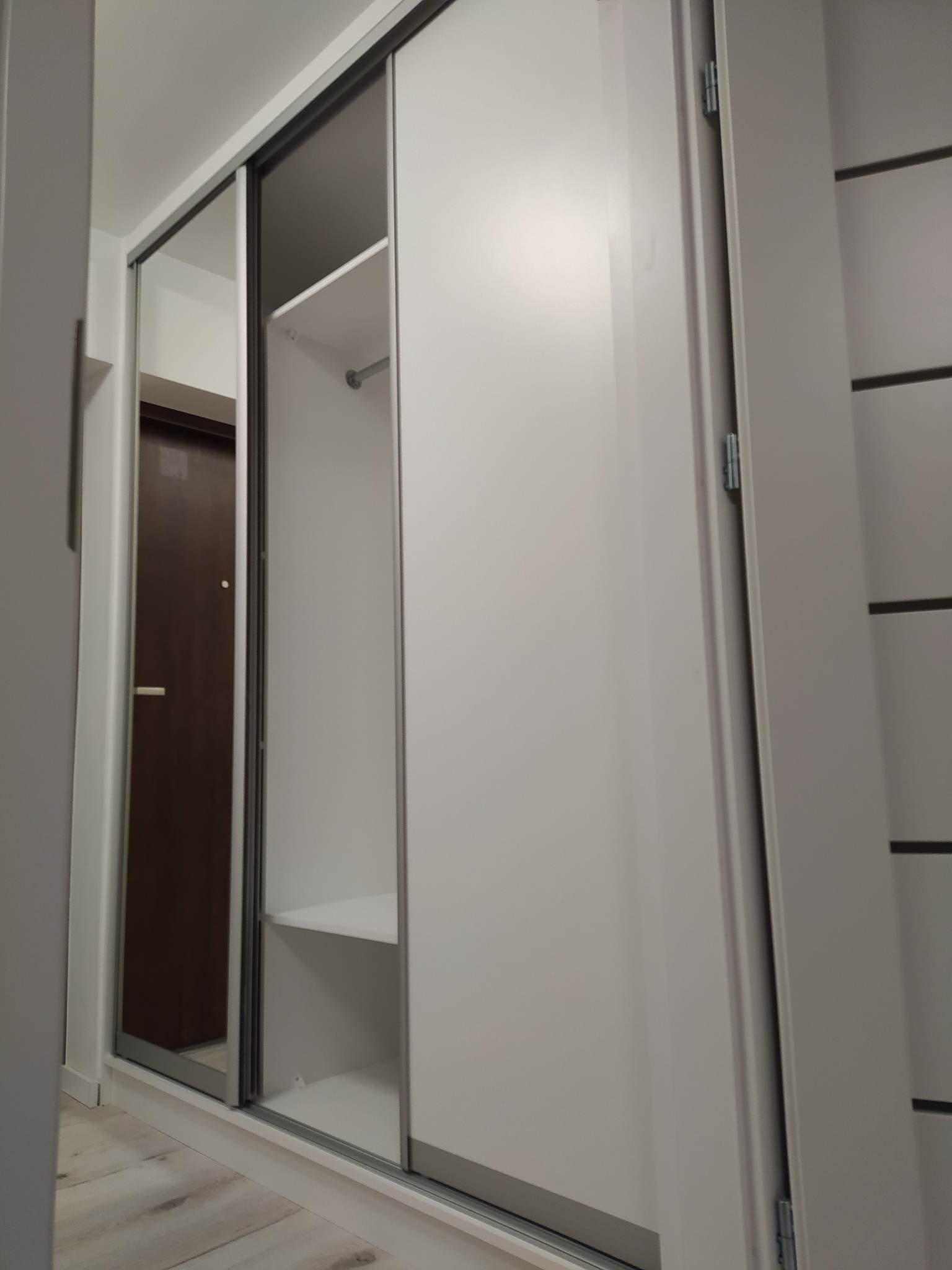 Biała szafa wnękowa z przesuwnymi drzwiami lustrzanymi, widoczne półki i drążek, minimalistyczny design.