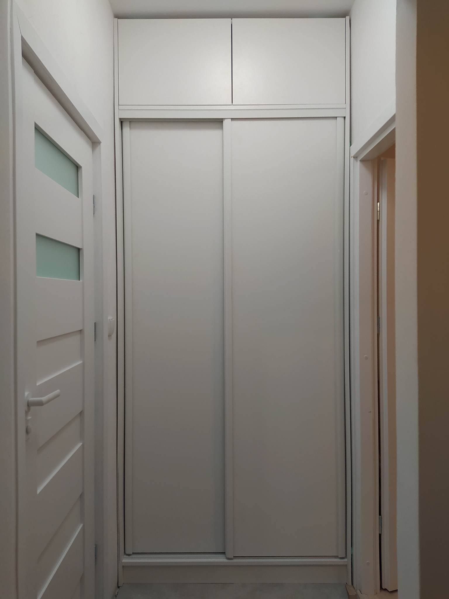 Wąski korytarz z zabudowaną szafą wnękową z przesuwnymi drzwiami w kolorze białym, sięgającą sufitu, obok widoczne drzwi pokojowe z przeszkleniami.