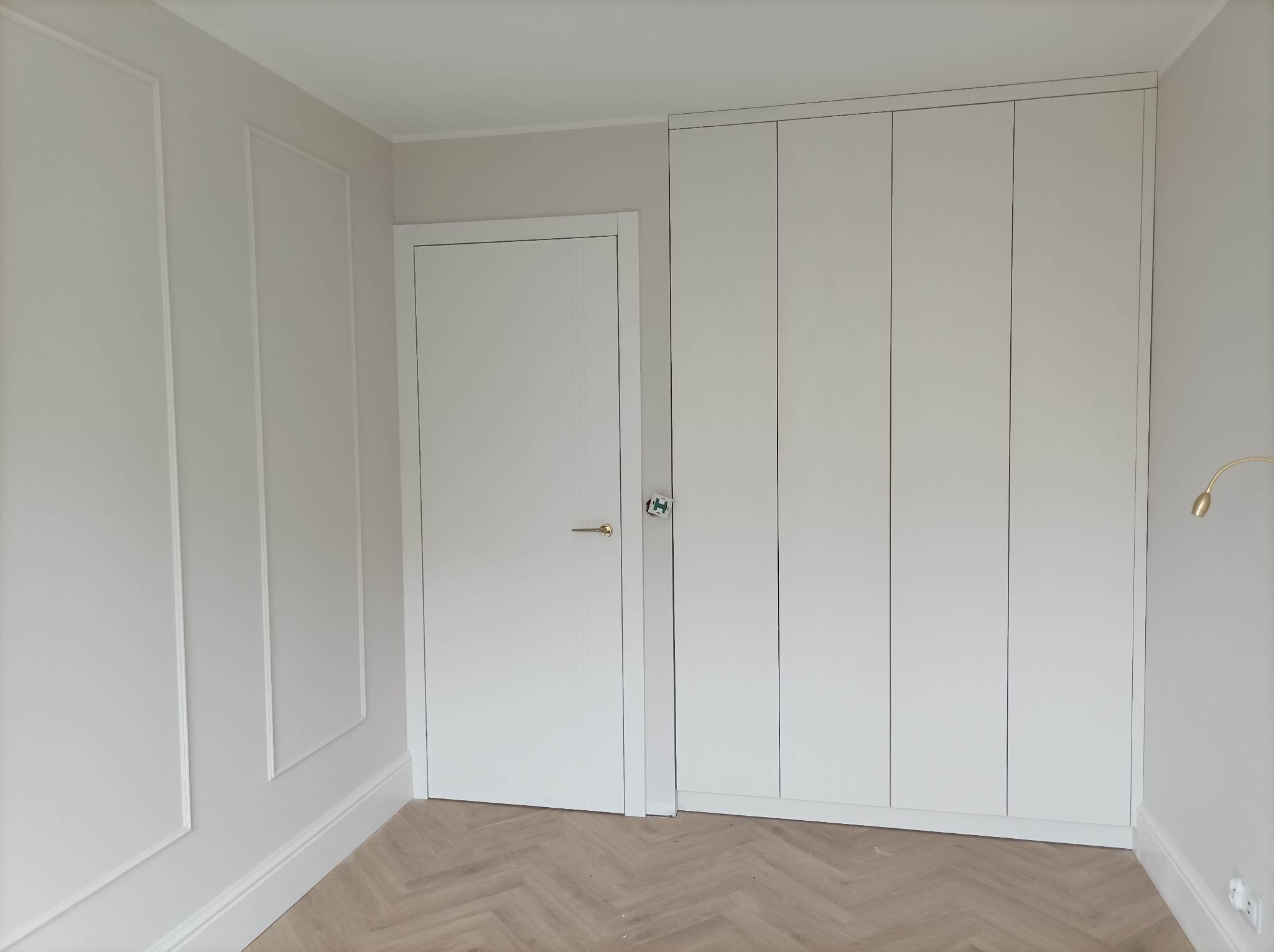 Minimalistyczne wnętrze w odcieniach bieli: zabudowana szafa z czterema frontami, drzwi wewnętrzne z prostą klamką i dekoracyjna listwa ścienna na tle parkietu ułożonego w jodełkę.