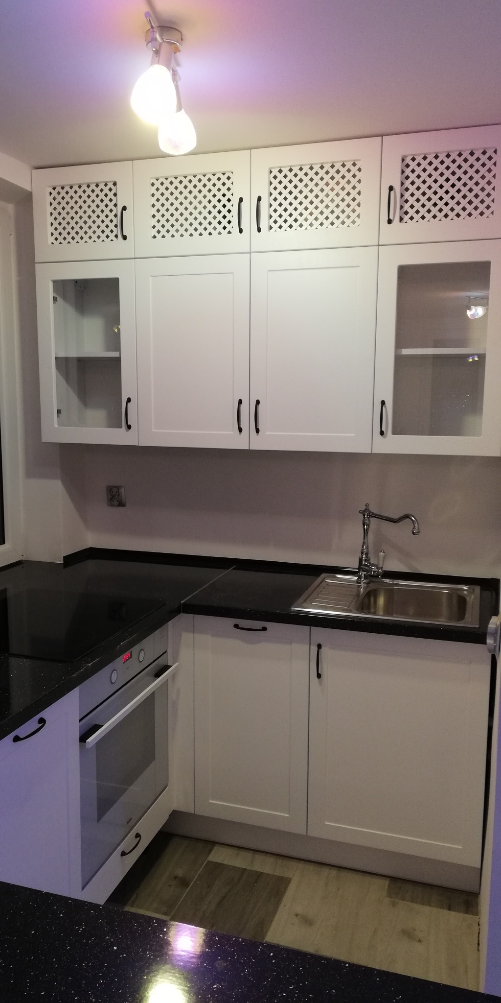 Nowoczesna, biała zabudowa kuchenna z czarnym, błyszczącym blatem w Gdańsku. Górne szafki z ażurowymi i przeszklonymi frontami, czarne uchwyty, oświetlenie punktowe.