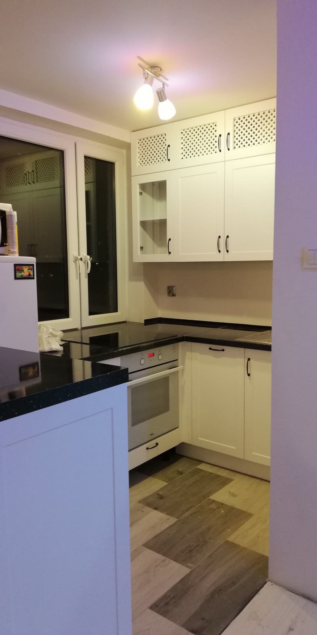 Biała kuchnia z czarnym blatem, górne szafki z ażurowymi frontami, oświetlenie punktowe, podłoga w odcieniach brązu ułożona w jodełkę.